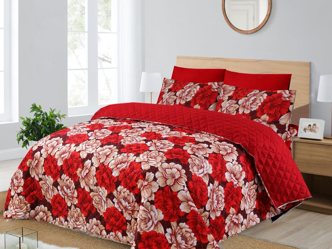 Red floral quilt set on a bed in a bedroom with a window and lamp.