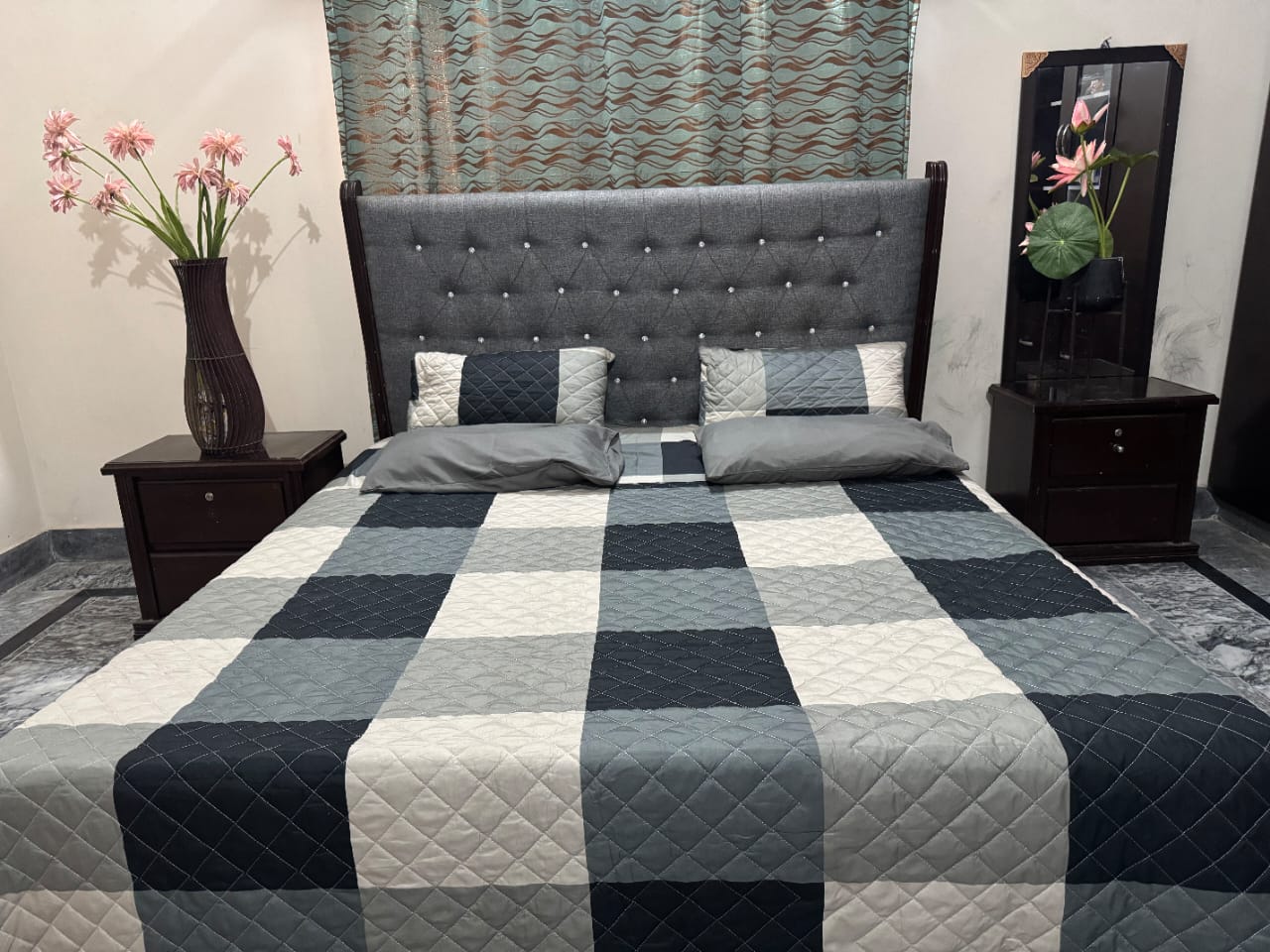 Bedroom with a bed featuring a gray checkered comforter, two nightstands with vases, and a patterned rug.